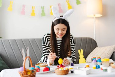 Evde paskalya yumurtası boyayan, tavşan kulaklığı takan, renkli süslemelerle çevrili ve Paskalya kutlamalarının neşeli atmosferinin tadını çıkaran mutlu genç bir kız.