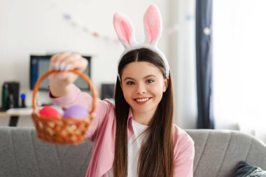 Tavşan kulaklığı takan gülümseyen genç bir kız Paskalya kutlaması sırasında evdeki kanepede otururken renkli paskalya yumurtalarıyla dolu sepeti gösteriyor. Doğu konsepti