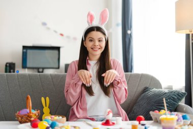 Tavşan kulaklığı takan genç bir kız, rahat bir koltukta oturuyor, rengarenk paskalya yumurtalarını süslerken gururla evdeki boyalı tırnaklarını sergiliyor. Doğu konsepti
