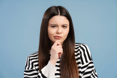 Bir karar verirken çenesine dokunan düşünceli genç bir kızın stüdyo portresi, bir çözüm düşünürken ya da parlak mavi bir arkaplan karşısında bir sorunu çözerken.