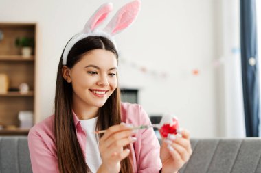 Genç kız evde paskalya yumurtası boyuyor, tavşan kulaklığı takıyor, gülümsüyor ve Paskalya kutlaması için hazırlanıyor, neşeli atmosferin tadını çıkarıyor. Kutlama konsepti