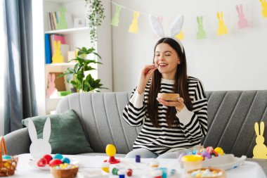 Tavşan kulaklı mutlu genç kız kanepede oturuyor, paskalya yumurtası boyuyor ve tatlı yiyor, evde şenlik hazırlıklarının tadını çıkarıyor. Kutlama konsepti
