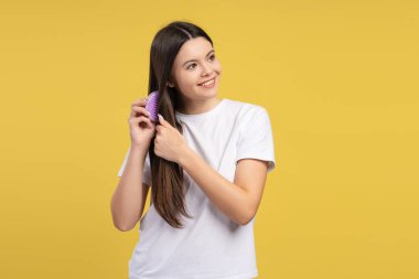 Gülümseyen genç bir kız, uzun kahverengi saçlarını mor bir tarakla tarıyor, canlı sarı arka plana bakıyor, saç bakımının ve güzelliğin özünü yakalıyor. Reklam kavramı