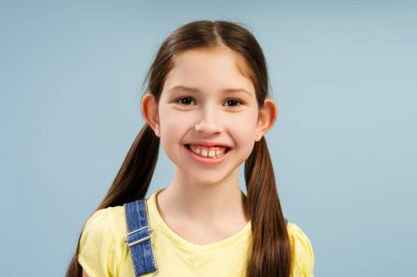 Saçı örgülü, sarı tişört ve kot tulumlu neşeli genç kızın stüdyo portresi, açık mavi bir arka plana geniş bir gülümseme, mutluluğu ve gençlik enerjisini aktarma, manken pozu