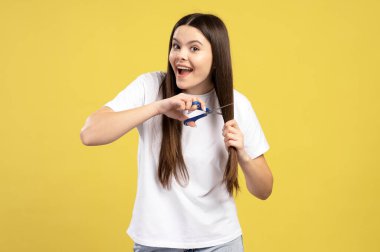 Uzun kahverengi saçlı tutkulu genç kız saçının bir kısmını tutuyor ve onu makasla kesiyor, canlı sarı bir arka planda heyecanı ifade ediyor. Saç bakımı konsepti