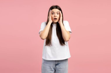 Uzun kahverengi saçlı, beyaz tişört ve kot pantolon giyen, yüzünü pembe arka plana karşı şaşkınlıkla kapatan şaşırmış genç bir kız. Alışveriş konsepti