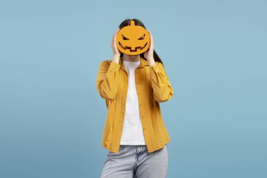 Yüzünün önünde cadılar bayramı balkabağı tutan genç bir kızın stüdyo portresi, kimliğini canlı bir mavi arka planda gizliyor, ürkütücü sezonu kutluyor. Kutlama konsepti