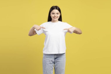 Gülümseyen kız işaret parmaklarıyla boş beyaz tişörtünü gösterirken aynı zamanda canlı sarı arka planda duruyor, özel tasarım ve mesajlar için yer sunuyor. Alışveriş, reklam konsepti.