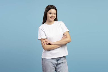 Kollarını çapraz bağlamış neşeli genç bir kızın canlı mavi bir arka plana karşı duran, kendine güveni ve olumlu bir tutum sergileyen bir stüdyo görüntüsü. Reklam kavramı