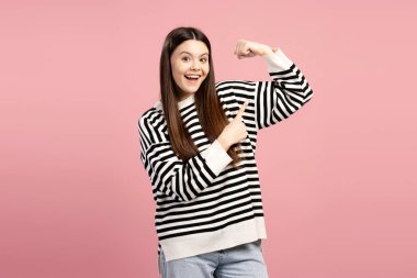 Neşeli bir genç kızın kaslarını esnetip parmağıyla göstererek pembe bir arka planda gücünü ve kararlılığını gösteren bir stüdyo fotoğrafı. Reklam kavramı