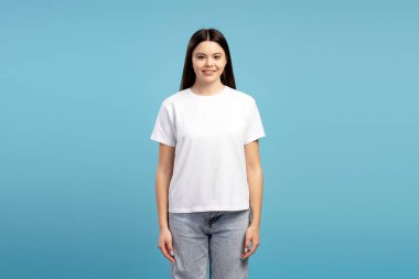 Beyaz tişört ve kot pantolon giyen neşeli genç bir kızın, canlı mavi bir fonda poz verirken, tasarım veya kavramları sergilemek için temiz ve modern bir estetik yaratırken,