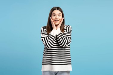 Uzun kahverengi saçlı, çizgili kazak giyen, elleriyle yüzüne dokunan ve mavi arka planda şaşkınlığını ifade eden şaşırmış genç bir kızın stüdyo portresi.