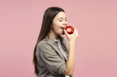 Uzun kahverengi saçlı, rahat bir gömlek giyen, canlı pembe bir zemine karşı kıtır kıtır kırmızı bir elmanın tadına bakan sağlıklı beslenme alışkanlıklarını teşvik eden genç bir kızın yan görüntüsü. Yiyecek konsepti