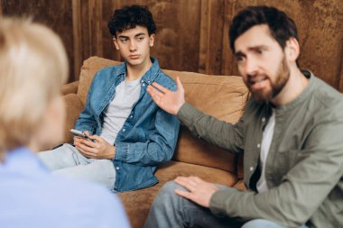 Aile terapisi sırasında üzgün baba ve genç oğlunun portresi, kadın psikoterapistle konuşmak. Aile sorunları, gençlik sorunları, terapist, tavsiye, yardım kavramı