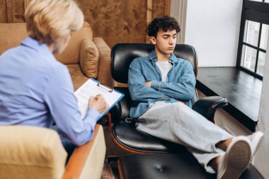 Terapi seansı sırasında genç çocuğa teşhis koyarken not defterine notlar yazan kadın terapist. Ruh sağlığı konsepti