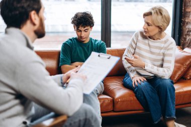 Profesyonel erkek psikolog notlar yazıyor, modern ofiste annesi ve ergen oğluyla konuşuyor. Danışmanlık terapisi. Psikoloji tedavisi