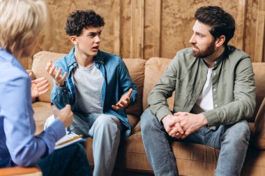 Genç bir çocuk kanepede otururken babasına ve psikoterapiste açıklama yapıyor. Aile sorunları, gençlik sorunları, terapist, tavsiye, yardım kavramı