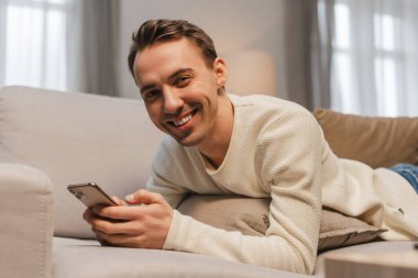 Yakışıklı, mutlu bir adam cep telefonu tutuyor, mobil uygulama kullanıyor, evdeki rahat kanepede uzanıyor. Çevrimiçi teknoloji, sosyal medya kavramı