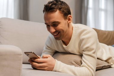 Gülümseyen, cep telefonu tutan, mobil uygulama kullanan, evdeki rahat kanepede uzanan mutlu bir adam. Çevrimiçi teknoloji, sosyal medya kavramı