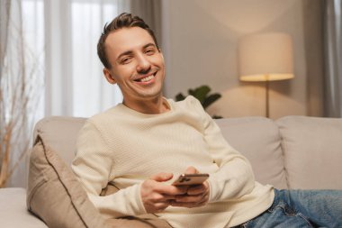 Evdeki rahat koltukta dinlenirken cep telefonu kullanan gülümseyen adam, online iletişimin ya da kameraya bakarak sosyal medyanın tadını çıkarıyor. Çevrimiçi teknoloji kavramı