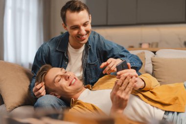 Evdeki mobil uygulamayı kullanarak koltukta rahat rahat oturan eşcinsel çift, oturma odasında rahat bir kanepede oturuyor. İlişki, lgbt kavramı