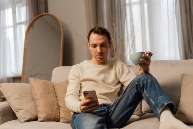 Pozitif, çekici bir adam rahat koltukta dinleniyor, akıllı telefon kullanıyor ve evde çay içiyor. Sosyal medya, sabah rutini kavramı