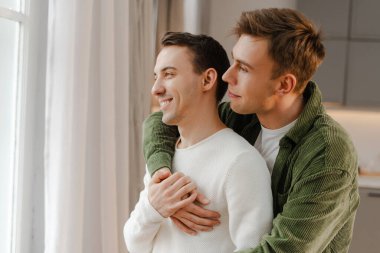 Romantik eşcinsel çift kucaklaşıyorlar ve pencereden dışarı bakıyorlar, evde birlikte sakin bir anın tadını çıkarıyorlar. Sevgi. ilişki kavramı