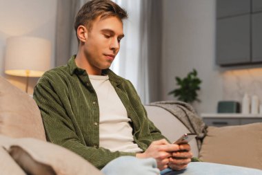 Evdeki rahat koltukta dinlenirken cep telefonu uygulaması kullanan genç bir adam. Çevrimiçi alışveriş kavramı