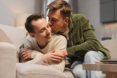 Mutlu eşcinsel çift, modern oturma odasındaki kanepede sarılıp öpüşen iki çekici erkek. İlişki, aşk kavramı