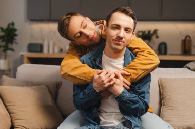Modern oturma odasında, evdeki kanepede kucaklaşan mutlu eşcinsel çift. Birlikte zaman geçiren gülümseyen adamlar. İlişki, aşk kavramı