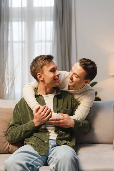 Happy gay couple, smiling two men embracing on comfortable sofa at home in living room. Love, relationship concept