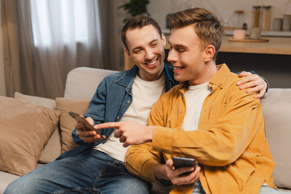 Two cheerful men are showing each other something on their smartphones while sitting on a sofa at home. Gay couple text message, hugging together. Browsing concept