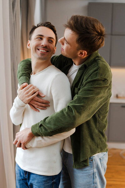 Two men hugging each other in their apartment, smiling and looking at each other standing in modern living room. Love, relationship concept