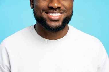 Kapalı stüdyo portresi, beyaz kazak giyen, sakallı, gülümseyen bir adamı, parlak mavi arka planda kusursuz beyaz dişlerini ön plana çıkarıyor. Diş sağlığı konsepti