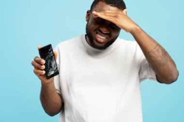 Hüsrana uğramış bir adam kırık bir akıllı telefonu kırık bir dokunmatik ekranla tutuyor ve yüzünü eliyle kapatıyor. Alet arızasından muzdarip, mavi stüdyo arka planında poz veriyor. Bozuk teknik kavram