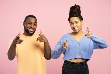 Gülümseyen Afro-Amerikan çift kamerayı işaret ediyor, özgüvenlerini dile getiriyorlar ve sizi seçiyorlar, pembe stüdyo arka planında birlikte poz veriyorlar,