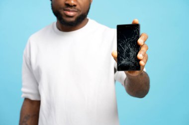Mavi arka planda çatlak bir ekranla akıllı telefonu elinde tutan bir adamın yakın plan çekimi, cep telefonu kazaları ve tamir veya yenileme ihtiyacını vurguluyor