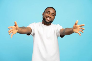 Beyaz tişört giyen genç, gülümseyen Afrikalı Amerikalı adam kollarını açıyor ve gülümsüyor, sarılıyor, mavi bir arka planda soyutlanmış, sevgi ve davetiye sunuyor. Hoş geldin konsepti