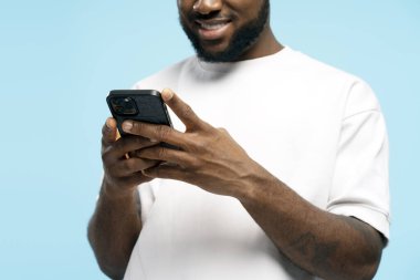 Gülümseyen Afrikalı genç bir adam cep telefonu kullanıyor, beyaz tişört giyiyor, mavi arka planda izole edilmiş, mesaj yazıyor ya da sosyal medyada geziniyor. Çevrimiçi iletişim kavramı