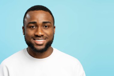 Beyaz tişört giyen, mavi arka plana özgüven ve pozitiflik saçan neşeli Afro-Amerikan bir adamın stüdyo portresi. Reklam kavramı