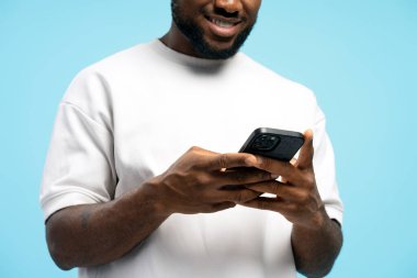 Elinde cep telefonu olan gülümseyen Afrikalı Amerikalı bir adam mavi arka planda mobil uygulama kullanıyor, ellerine odaklanıyor. Çevrimiçi teknoloji kavramı