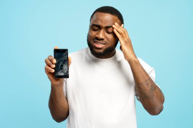 Üzgün Afrikalı Amerikalı bir adam hayal kırıklığına uğramış bir ifadeyle bozuk bir akıllı telefon dokunmatik ekranı gösteriyor, elleriyle kafasına dokunuyor, mavi arka planda duruyor. Çevrimiçi teknoloji kavramı