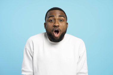Beyaz tişört giyen, ağzı açık şokta olan, şaşkın bir genç Afro-Amerikalı 'nın portresi, canlı mavi bir arka plana karşı şaşkınlık ifadesi sergiliyor, duygular kavramı