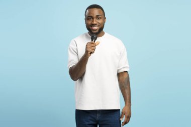 Kendinden emin genç Afrikalı Amerikalı bir adam mikrofon tutarak şarkı söylüyor, mavi stüdyo arka planında duruyor, günlük beyaz tişört giyiyor, gülümsüyor ve kameraya bakıyor. Reklam kavramı