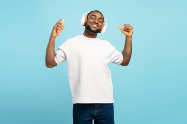 Kulaklıklı ve akıllı telefonlu mutlu Afrikalı Amerikalı adam müzik dinliyor, gözleri kapalı dans ediyor ve şarkı söylüyor, mavi stüdyo arka planında izole edilmiş. Eğlenceli konsept