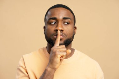 Sessiz bir jest yapan, sessizlik ya da gizlilik isteyen, işaret parmağını dudaklarına bastıran, bej arkaplana bakan ciddi bir siyahi adamın portresi. Sessizlik kavramı