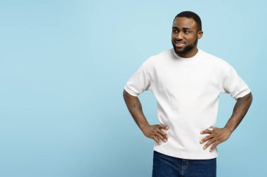 Genç, gülümseyen, elleri kalçasında, mavi arka planda, beyaz tişört ve kot pantolon giymiş, Afro-Amerikan bir adam. Reklam kavramı
