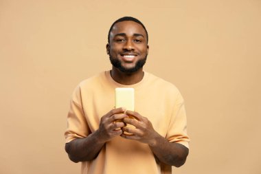 Neşeli genç yetişkin adam elinde boş ekranlı akıllı telefonu tutuyor ve bej arka planda gülümsüyor, yeni mobil uygulama veya modern teknolojinin reklamını yapıyor.