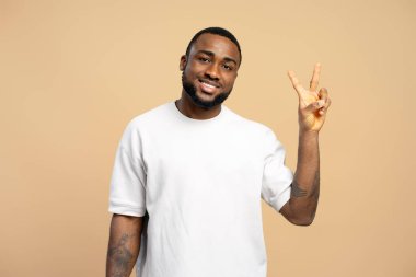 Neşeli, Afro-Amerikan bir gencin portresi zafer işaretini iki parmakla gösteriyor, bej renkli bir stüdyonun arka planında duruyor, başarısını ve mutluluğunu ifade ediyor.