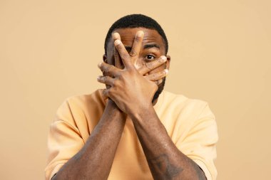 Tarafsız bir arkaplanda korku, utanç ya da kaygı hisseden, elleriyle yüzünü örten Afrikalı genç bir adamın stüdyo portresi.